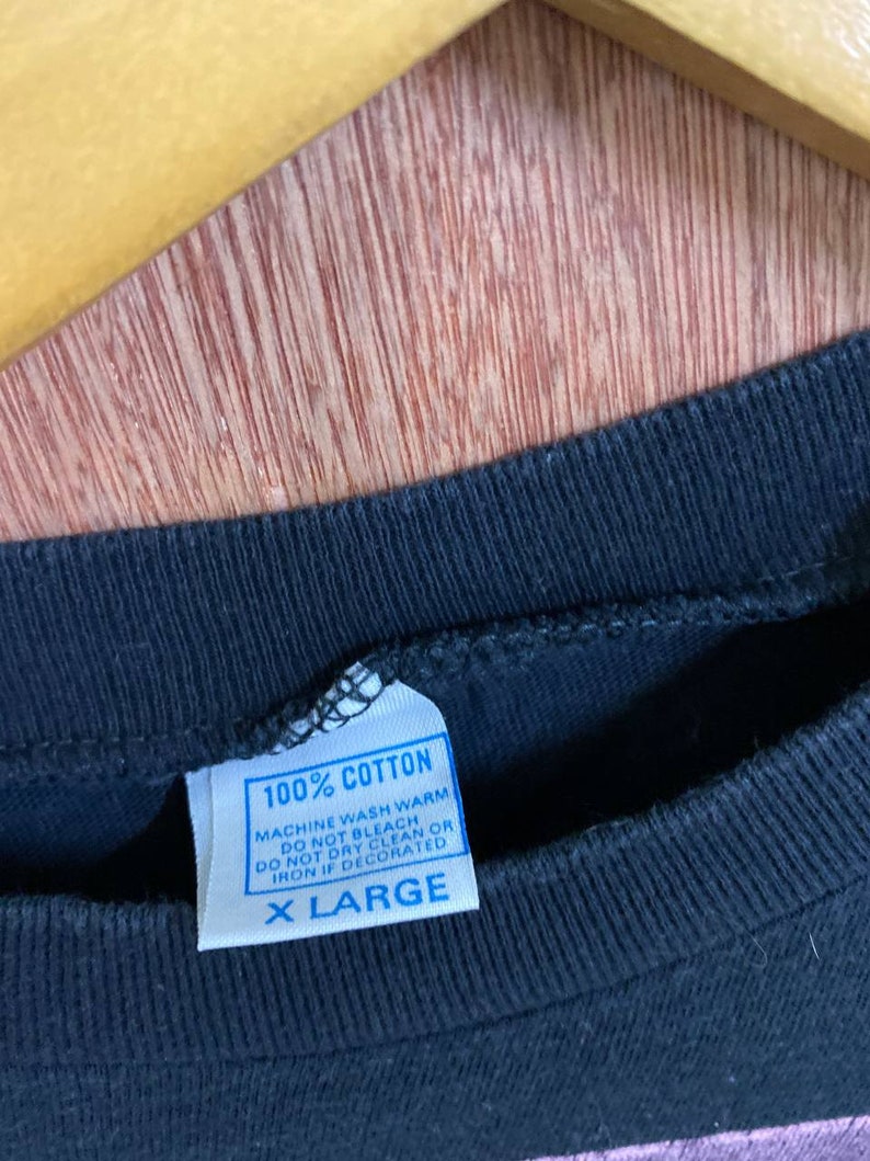 Puede incluir: Una camiseta negra con una etiqueta de cuidado azul que dice "100% Algod&oacute;n, Lavar a m&aacute;quina en caliente, No usar lej&iacute;a, No limpiar en seco o planchar si est&aacute; decorado, Talla XL."