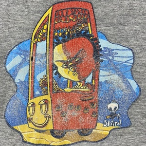K&ouml;nnte beinhalten: Ein Vintage-T-Shirt in Grau mit einem abgenutzten Grafikdruck eines Comic-Charakters, der ein rotes Auto mit einem Smiley-Gesicht f&auml;hrt. Das Auto hat ein gelbes Dach und R&auml;der. Der Charakter hat einen roten Kopf und tr&auml;gt ein gelbes Hemd. Der Text "Blind" ist in Wei&szlig; in der unteren rechten Ecke des Grafikdrucks gedruckt.