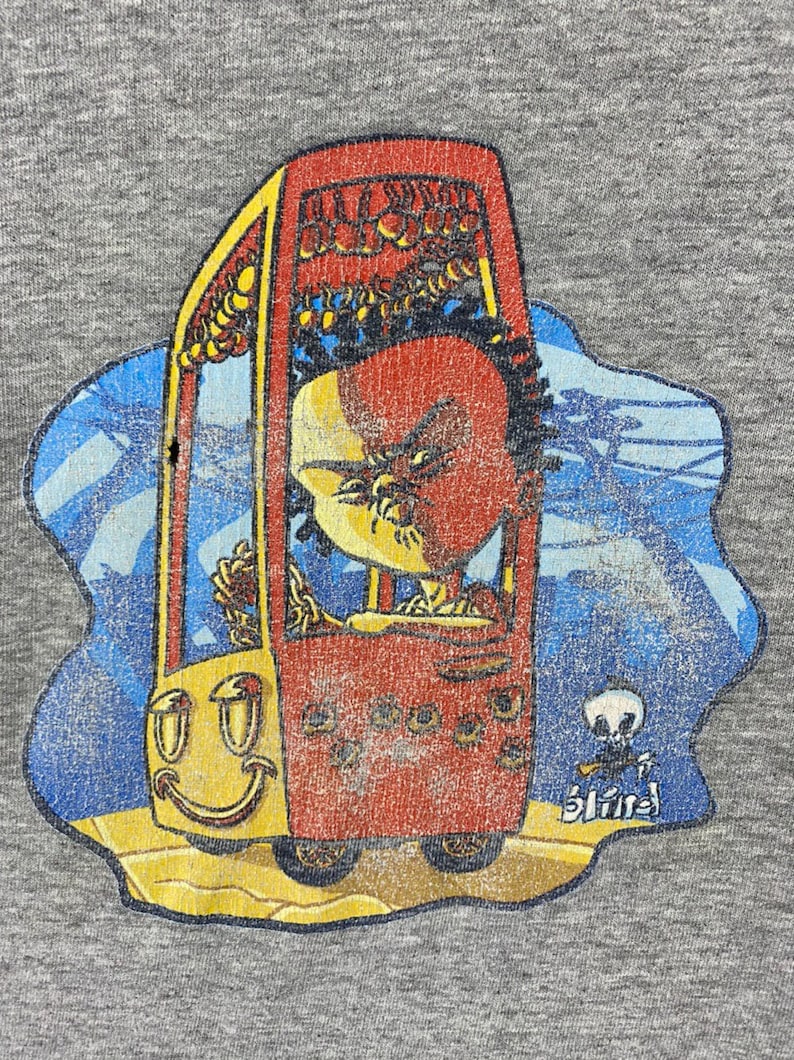 K&ouml;nnte beinhalten: Ein Vintage-T-Shirt in Grau mit einem abgenutzten Grafikdruck eines Comic-Charakters, der ein rotes Auto mit einem Smiley-Gesicht f&auml;hrt. Das Auto hat ein gelbes Dach und R&auml;der. Der Charakter hat einen roten Kopf und tr&auml;gt ein gelbes Hemd. Der Text "Blind" ist in Wei&szlig; in der unteren rechten Ecke des Grafikdrucks gedruckt.