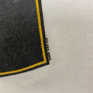 Pu&ograve; includere: Primo piano di una maglietta bianca con una grafica nera e gialla. La grafica &egrave; un quadrato con sfondo nero e bordo giallo. Il testo "Grateful Dead" &egrave; visibile nel bordo giallo.