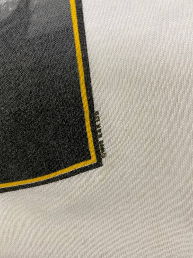 Pu&ograve; includere: Primo piano di una maglietta bianca con una grafica nera e gialla. La grafica &egrave; un quadrato con sfondo nero e bordo giallo. Il testo "Grateful Dead" &egrave; visibile nel bordo giallo.