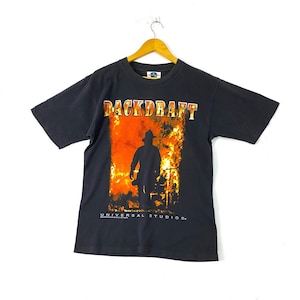 May include: Black t-shirt featuring a graphic print of a firefighter walking through flames. The text "BACKDRAFT" is printed in white above the graphic. The text "UNIVERSAL STUDIOS." is printed in white below the graphic.