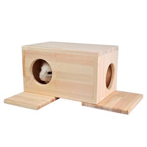 Può includere: Una casetta di legno per animali domestici con due ingressi e una rampa. La casetta è realizzata in legno naturale e ha un colore marrone chiaro. Un piccolo animale bianco spunta da uno degli ingressi.