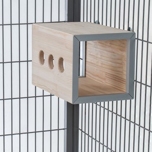 May include: A wooden cage accessory with a square opening and three round holes. The accessory is made of light wood with a grey trim and is attached to a grey metal cage.