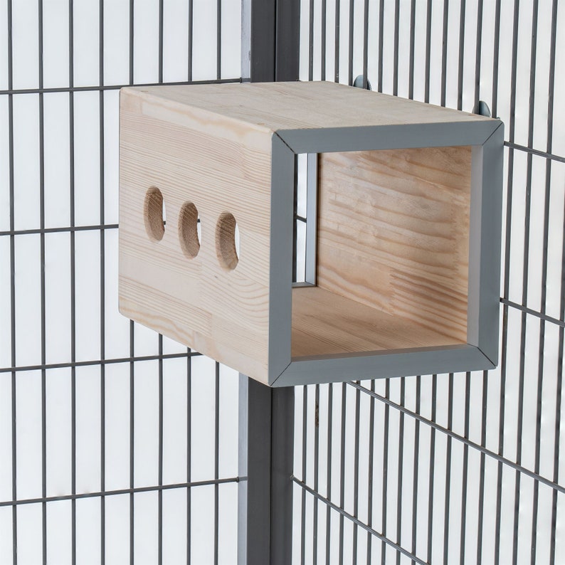 May include: A wooden cage accessory with a square opening and three round holes. The accessory is made of light wood with a grey trim and is attached to a grey metal cage.