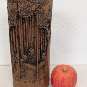 May include: A cylindrical, brown bamboo vase with intricate carved relief depicting a bamboo forest scene with figures. A red apple sits beside the vase. The vase is approximately 6 inches tall.