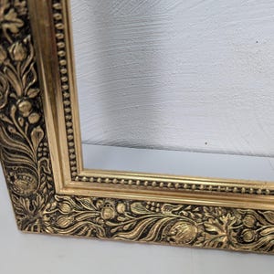 May include: A rectangular picture frame with a metallic gold finish and a dark recessed background. The frame features an ornate relief pattern of leaves, berries, and floral motifs along the border, complemented by a beaded inner edge detail.