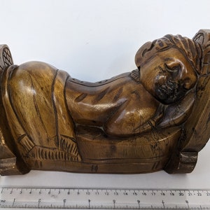 May include: A carved wooden sculpture of a sleeping figure reclining on a carved bed. The sculpture is made of dark brown wood and features detailed carvings of the figure's face, hair, and clothing. The bed has decorative armrests.