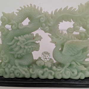 Può includere: Una scultura color giada raffigurante un drago e una fenice con una sfera di cristallo. Il drago è a sinistra e la fenice a destra. La scultura è montata su una base nera.