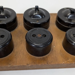 May include: A wooden board holds six vintage black electrical switches and outlets. Three switches have a lever, and three outlets have two holes. The items are arranged in two rows of three.