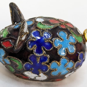 Puede incluir: Una pequeña figura de cerdo de cloisonné negro con un anillo de cola dorado. El cerdo está decorado con flores de esmalte de colores en tonos de azul, verde, rojo y blanco.
