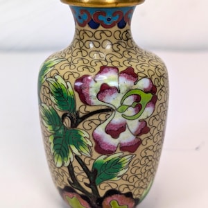 May include: A small cloisonné vase featuring a beige background with a repeating black wire scroll pattern. The vase is decorated with a large pink and white flower, green leaves, and a gold-toned metal rim at the top and bottom opening.