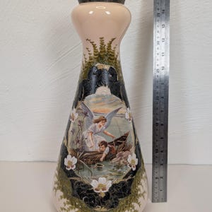 May include: A tall, cream-coloured vase with a dark metallic rim, featuring a central oval illustration of an angel and a child in a boat. The vase is decorated with green foliage patterns and white flowers. A metal ruler shows a height of 41 cm.