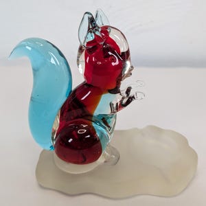 Op de afbeelding: Een kleurrijk glazen eekhoornbeeldje met een lichtblauwe staart en een rode body. De eekhoorn staat op een matglazen voet. Het beeldje is een decoratief item.