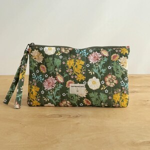 Op de afbeelding: Een rechthoekig gewatteerd tasje met rits in donkergroen met een veelkleurige bloemenprint van gele, roze en witte bloemen. Het tasje is voorzien van een bijpassend polsbandje en een klein wit rechthoekig label met THE PROJECT BAG.