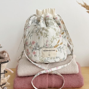 Puede incluir: Un bolso acolchado con cordón y estampado floral en tonos suaves de rosa, verde y amarillo sobre un fondo crema. El bolso tiene una etiqueta de tela en la parte delantera que dice THE PROJECT BAG by 314 e incluye dos correas de tela a juego.