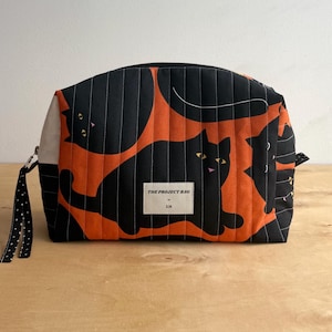 Op de afbeelding: Een gewatteerd stoffen tasje met een herhalend patroon van zwarte katten op een oranje achtergrond. Het tasje heeft een zwarte ritssluiting met een zwart-wit gestippeld treklipje en een wit rechthoekig label met de tekst THE PROJECT BAG by JLM.
