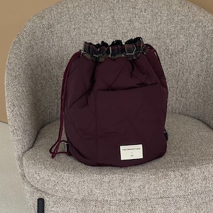 Op de afbeelding: Een diepbordeauxrode gewatteerde tas met trekkoordsluiting en een geruite stoffen rand bij de opening. De tas heeft bijpassende bordeauxrode trekkoorden en een klein wit rechthoekig label aan de voorzijde met de tekst THE PROJECT BAG en SW.