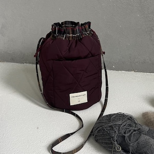 Op de afbeelding: Een gewatteerde bordeauxrode bucket bag met een trekkoordsluiting en een geruite voering. De tas heeft een lange geruite schouderband en een wit rechthoekig label aan de voorzijde met de tekst THE PROJECT BAG en een klein logo eronder.
