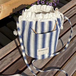 Op de afbeelding: Een blauw-wit gestreepte stoffen emmertas met een trekkoordsluiting en een geplooide bovenrand. De tas heeft een buitenvak met twee houten breinaalden en een rechthoekig label met de tekst THE PROJECT BAG by 3.14.