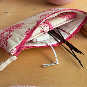 Op de afbeelding: Een gewatteerd etui met rits en een crème en felroze toile de jouy-patroon. Het open etui bevat een wit meetlint, diverse donkere metalen breinaalden en een crème notitieboek met een bijpassende stoffen riemsluiting.