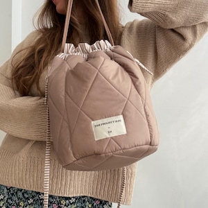 Pode incluir: Uma bolsa com cordão acolchoada cor de rosa com um padrão de diamante. A bolsa tem um interior listrado e uma alça a condizer. O texto "THE PROJECT BAG" está impresso numa etiqueta na frente da bolsa.