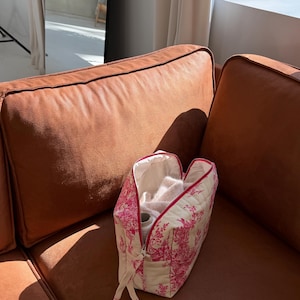 May include: A white and pink toiletry bag with a red zipper and trim, open to reveal contents. The bag is quilted and sits on a brown leather couch. The bag's design features a repeating pattern of pink floral and architectural elements.