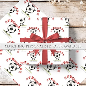 Puede incluir: Papel de regalo con un patr&oacute;n repetido de balones de f&uacute;tbol con bufandas, personalizado con el nombre "ALBIE" y el texto "Happy 4th Birthday". Una cinta y un lazo rojos envuelven el regalo. Tambi&eacute;n se ve el texto "MATCHING PERSONALISED PAPER AVAILABLE".
