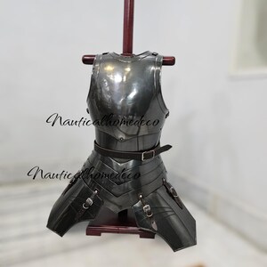 May include: A dark gray metal chest armor with attached leg protection, displayed on a wooden stand. The armor has a polished finish and is secured with leather straps and buckles. The text "Nauticalhomedeco" is visible.