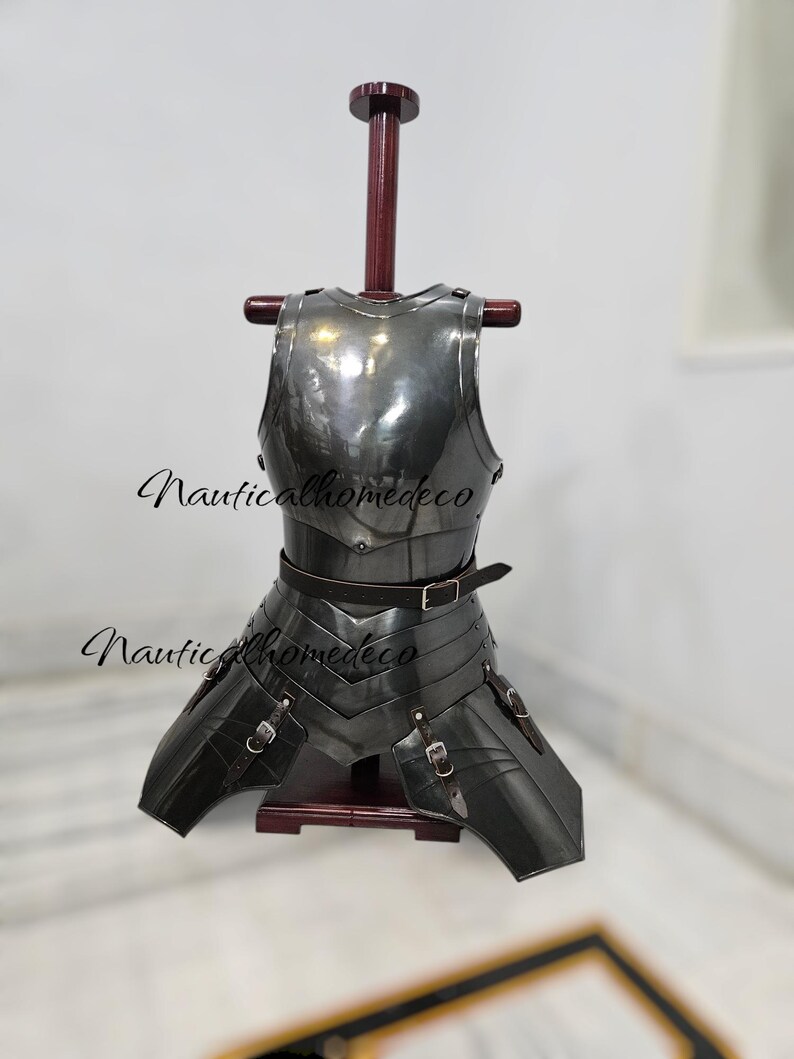 May include: A dark gray metal chest armor with attached leg protection, displayed on a wooden stand. The armor has a polished finish and is secured with leather straps and buckles. The text "Nauticalhomedeco" is visible.