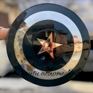 May include: A black and silver Captain America shield with a large, five-pointed star in the centre. The shield is made of metal and has a shiny finish.