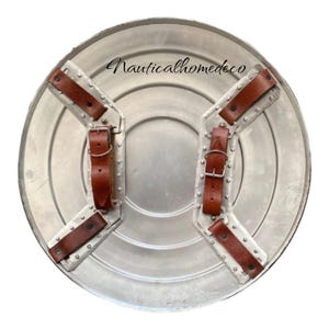 K&ouml;nnte beinhalten: Ein silberner Metallschild mit braunen Lederriemen und Nieten. Der Schild ist rund und hat eine leicht erh&ouml;hte Mitte. Der Text "Nauticalhomedeco" ist auf dem Schild aufgedruckt.