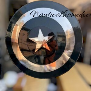May include: A black and silver Captain America shield with a white star in the centre. The shield is made of metal and has a shiny finish. The text "Nauticalhomedecor" is visible in the top right corner of the image.