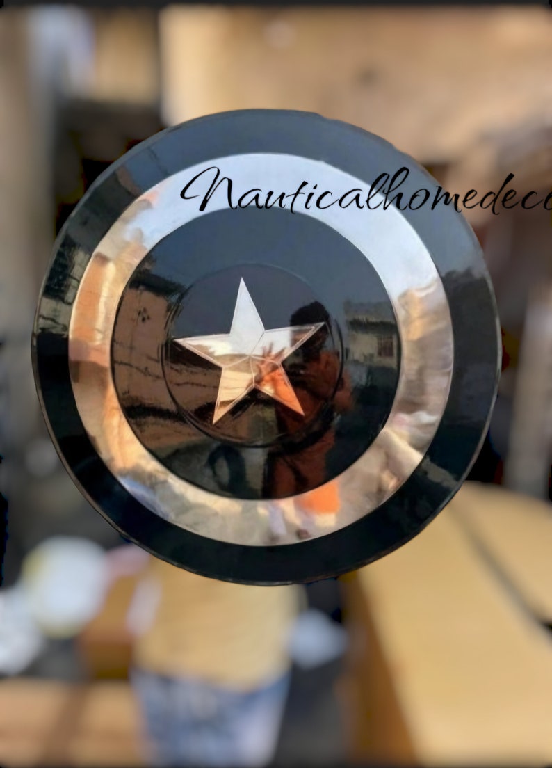 May include: A black and silver Captain America shield with a white star in the centre. The shield is made of metal and has a shiny finish. The text "Nauticalhomedecor" is visible in the top right corner of the image.