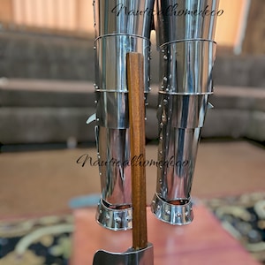 May include: A pair of silver metal leg armor with a wooden handle axe attached. The armor is made of multiple sections and has a shiny finish. The axe has a silver blade and a brown wooden handle.