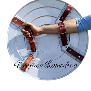 May include: A white metal shield with a red star in the centre. The shield has three brown leather straps with metal buckles. The shield is being held by a person's arm.