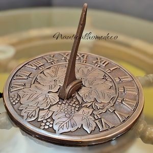 May include: A bronze sundial with Roman numeral markings and a grapevine design. The sundial has a pointed gnomon and is set on a glass surface.
