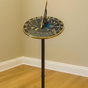 May include: A bronze sundial with a circular face and a vertical gnomon. The face features Roman numerals and symbols, mounted on a black metal stand. The sundial is a decorative item for outdoor spaces.