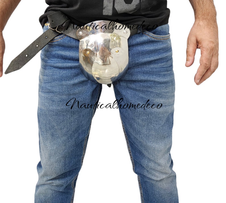 May include: A polished metal codpiece with a bear-like design, attached to a black leather belt. The item is worn over blue jeans. The text "Nauticalhomedeco" is visible on the image.