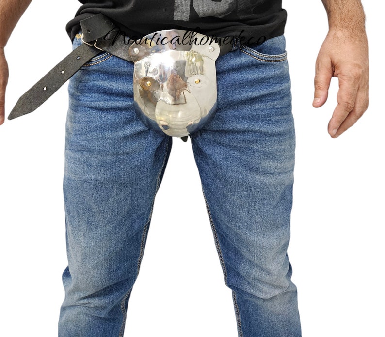 May include: A polished metal codpiece with a bear-like face design, attached to a black leather belt. The belt is worn over blue denim jeans. The image is set against a white background.