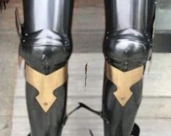 May include: A pair of black and gold medieval leg armor. The armor is made of metal and has a pointed toe. The armor is worn on a pair of legs.