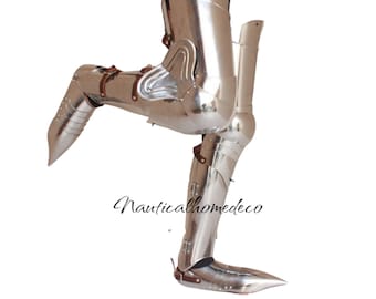 May include: Polished silver metal leg armor with articulated joints and leather straps. The armor is designed to protect the leg from the knee down to the foot. The armor is a historical item, likely from the medieval period.