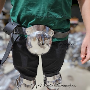 May include: A set of silver-coloured medieval armour for the legs. The armour includes knee pads and a metal codpiece that is attached to a black leather belt.