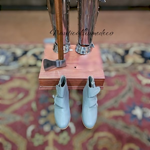 May include: A pair of gray leather boots with buckles hang from a wooden shelf. A small axe with a wooden handle is on the shelf. The shelf is attached to a pair of silver metal leg armor.