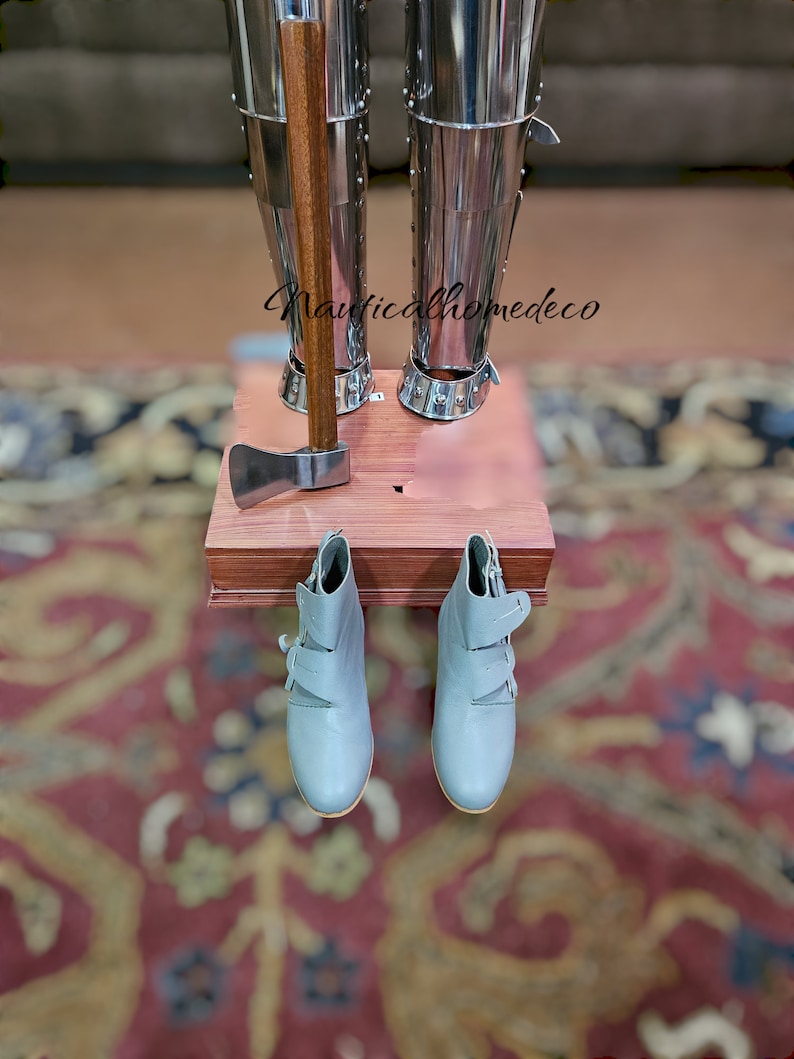 May include: A pair of gray leather boots with buckles hang from a wooden shelf. A small axe with a wooden handle is on the shelf. The shelf is attached to a pair of silver metal leg armor.
