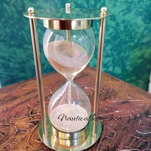 Puede incluir: Un reloj de arena de latón con un bulbo de vidrio transparente. El reloj de arena está lleno de arena y tiene una base y una parte superior de latón.