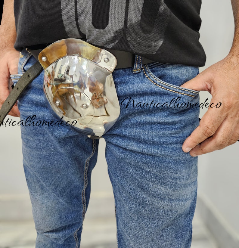 May include: A polished metal hip flask with a curved design, attached to a black leather belt. The flask has a shiny, reflective surface and is secured with rivets. The person is wearing blue jeans and a black shirt.