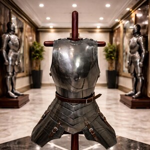 May include: A polished steel breastplate and faulds armor set displayed on a wooden stand. The armor features a brown leather belt and straps with buckles. Two full suits of armor stand in the background.