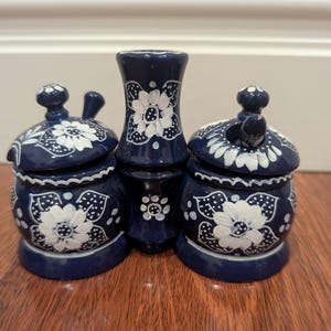 May include: A trio of dark blue ceramic items featuring white floral designs. The set comprises two lidded jars and a tall vase. The hand-painted floral pattern adds a touch of elegance. Ideal for kitchen display or storage.