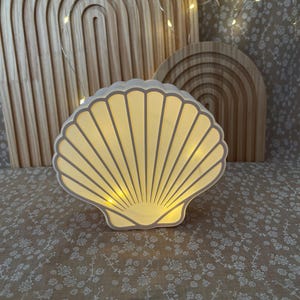 May include: A seashell-shaped lamp emitting a warm glow. The lamp is white with grey detailing and is placed on a floral patterned surface. Two wooden arch decorations are in the background, with fairy lights.